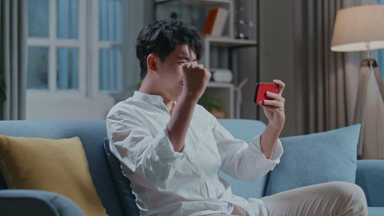 Asian Man Celebrating Winning Game On Smartphone While Sitting On Sofa In The Living Room