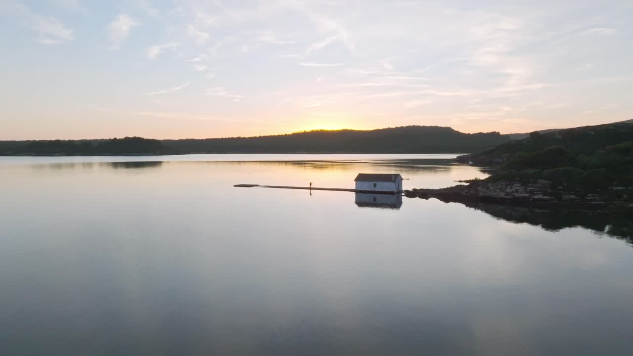 드론은 메노르카에 있는 포넬스 만의 반사된 물 위에서 낮게 날아다니며 스페인 여행 해안 목적지, 일몰, 다채로운 지평선 배경, 공중 전망