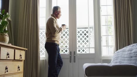 Happy-caucasian-man-looking-through-window-and-drinking-coffee-in-bedroom