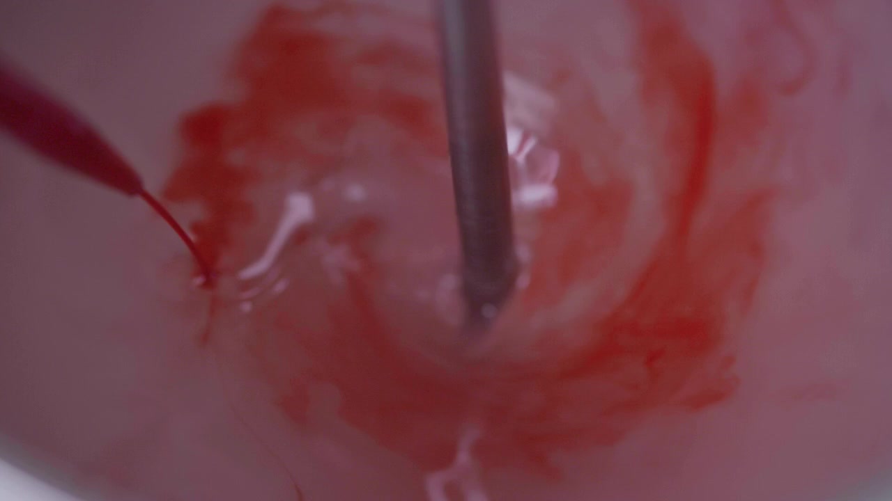 Nærbilde som viser en rød væske som helles med en pipette på en lys rosa base, mens en metallstang kontinuerlig roterer for å integrere begge komponentene.