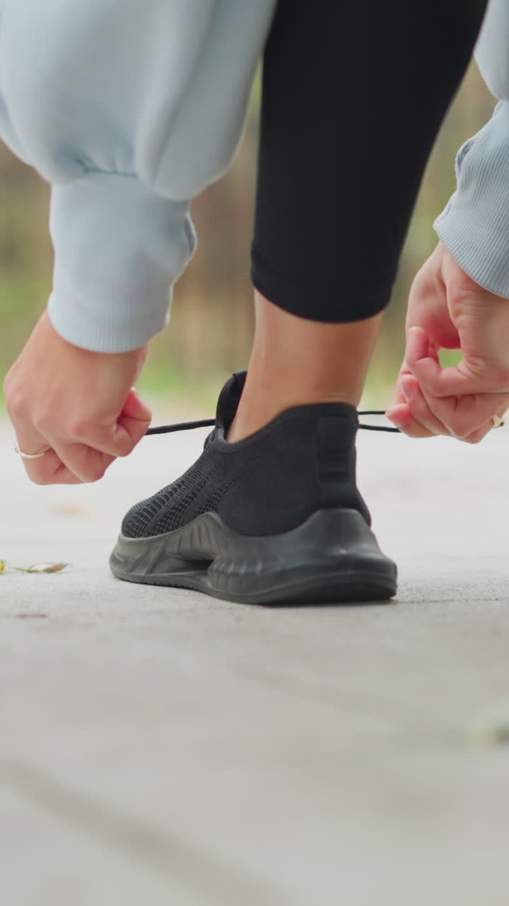 Back view of lady in black leggings and blue sweater tying her left shoe lace on sidewalk in park, preparing for a workout or run outdoors, focusing on active lifestyle and fitness