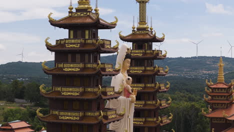 Aerial-view-of-Buddhist-temple-with-statue-in-Vietnam-and-rotating-wind-turbines-in-hill-in-background