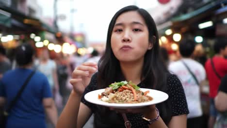 A young woman tastes flavorful street food at a bustling night market in Thailand. The lively environment features colorful stalls and a variety of delightful dishes being served.