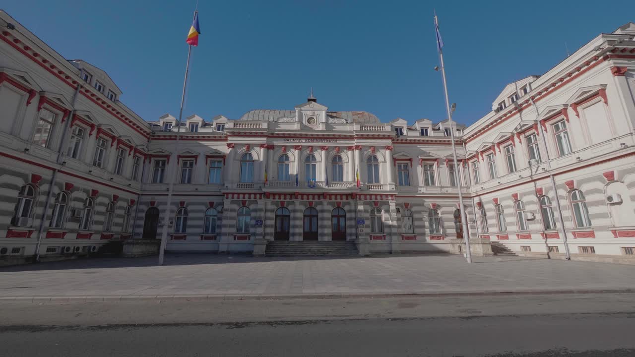 A cinematic, slow-motion dolly shot moves across a public square towards the historic Administrative Palace in Bacau, Romania, a grand neoclassical government building