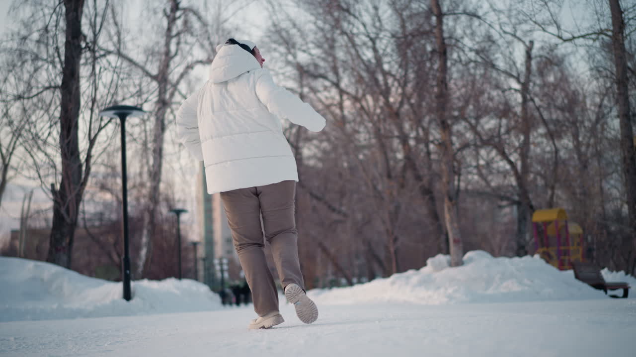 Turning dancer spins energetically in snowy park among bare trees at dusk wearing white puffer coat beanie and headphones conveying joyful winter rhythm and playful movement under pastel sky