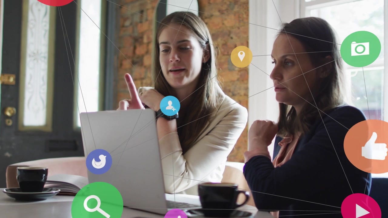 Due colleghe collaborano al laptop, con icone social che si attivano dal dialogo e mostrano dati di marketing.