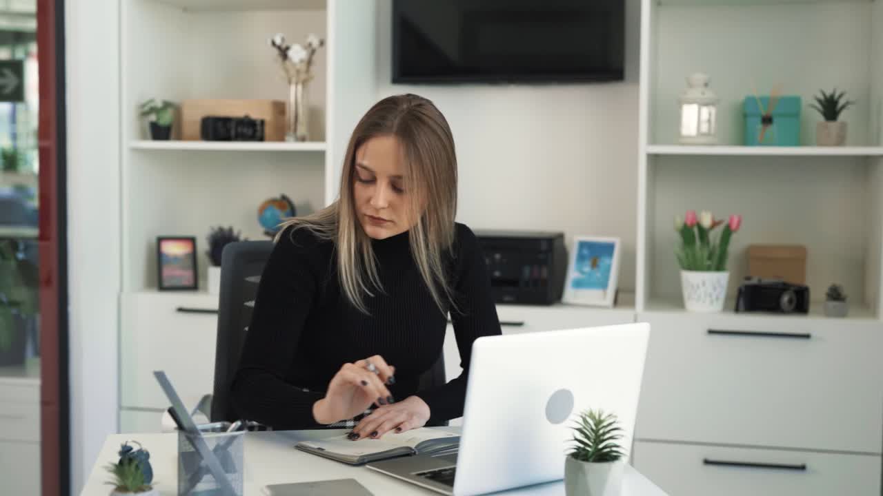 The young woman is working on a laptop in the office, taking notes in a notebook, and looking at her phone, she is very focused and attentive at work