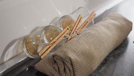 three glass cups arranged on burlap sack, shadows of person reflected on tiled wall while moving, artistic setup for candle making with materials carefully placed