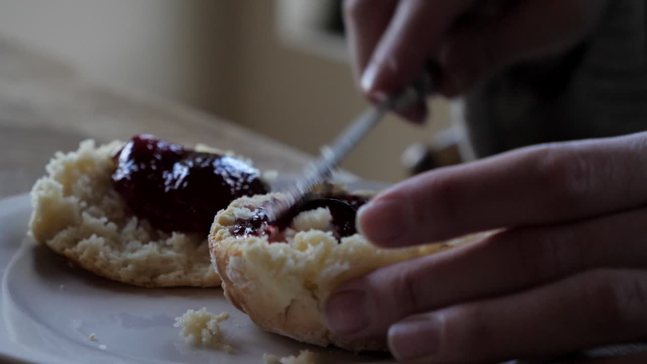 Up close shot of female hand spreading dollop of jam across two freshly baked traditional homemade scones on white plate and timber table for afternoon tea