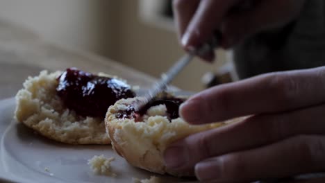 Primer-Plano-De-La-Mano-Femenina-Extendiendo-Una-Cucharada-De-Mermelada-A-Través-De-Dos-Bollos-Caseros-Tradicionales-Recién-Horneados-En-Un-Plato-Blanco-Y-Una-Mesa-De-Madera-Para-El-Té-De-La-Tarde