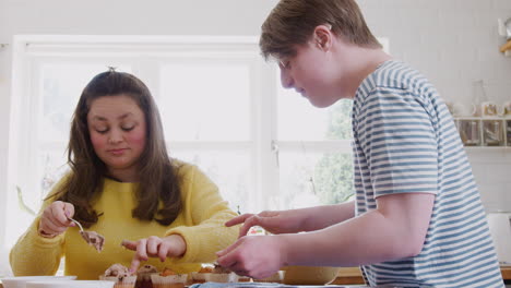 young downs syndrome couple decorating homemade cupcakes with icing in kitchen at home