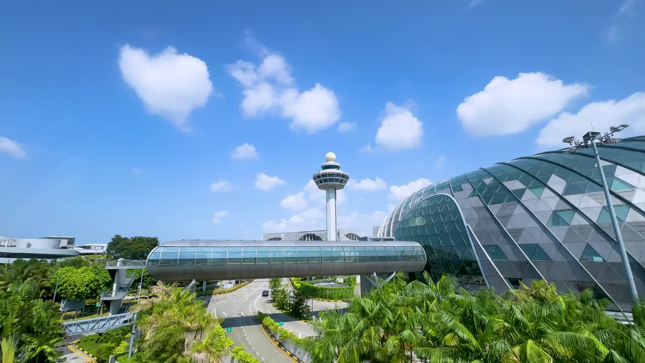 Smooth daytime aerial pan reveals a futuristic glass terminal, lush greenery, and a prominent control tower under clear blue skies with scattered clouds