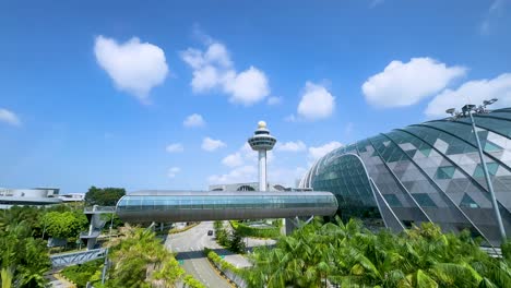 smooth daytime aerial pan reveals a futuristic glass terminal, lush greenery, and a prominent control tower under clear blue skies with scattered clouds
