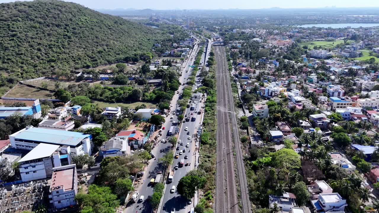 Aerial view of a major Chennai–Trichy highway (GST Road) and railway tracks in Chennai, flanked by a green hill on one side and urban development on the other, under a clear sky.