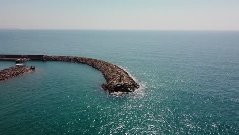 Un-Rompeolas-Curvo-En-Garraf,-Costa-Barcelona-En-Un-Día-Soleado,-Mar-Resplandeciente,-Vista-Aérea