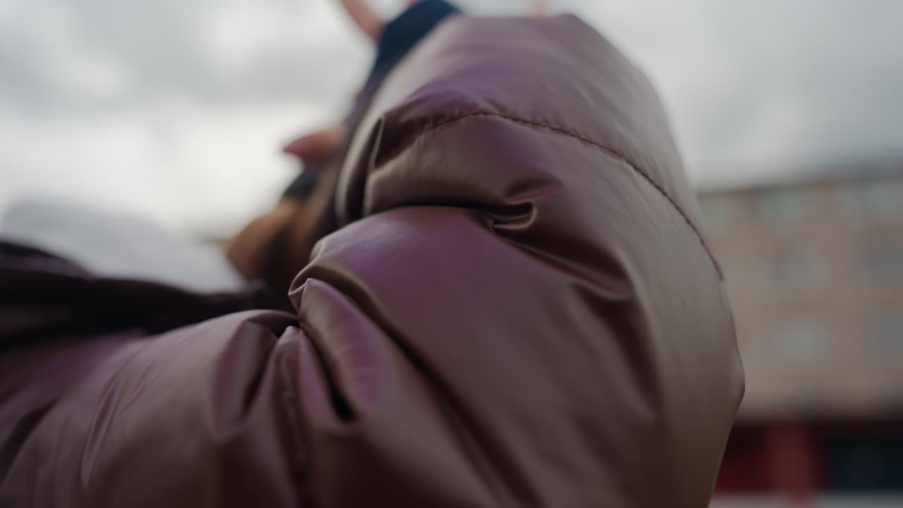 Mujer ajustándose el sombrero; mujer tirando suavemente de su gorro de punto hacia abajo; mujer curiosa con abrigo acolchado ajustándose el gorro al aire libre; joven con abrigo mullido y nubes de fondo bajándose activamente el sombrero