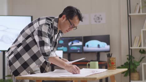 side view of asian male organizing papers while working about car design sketch on table in the studio with tv and computers display 3d electric car model