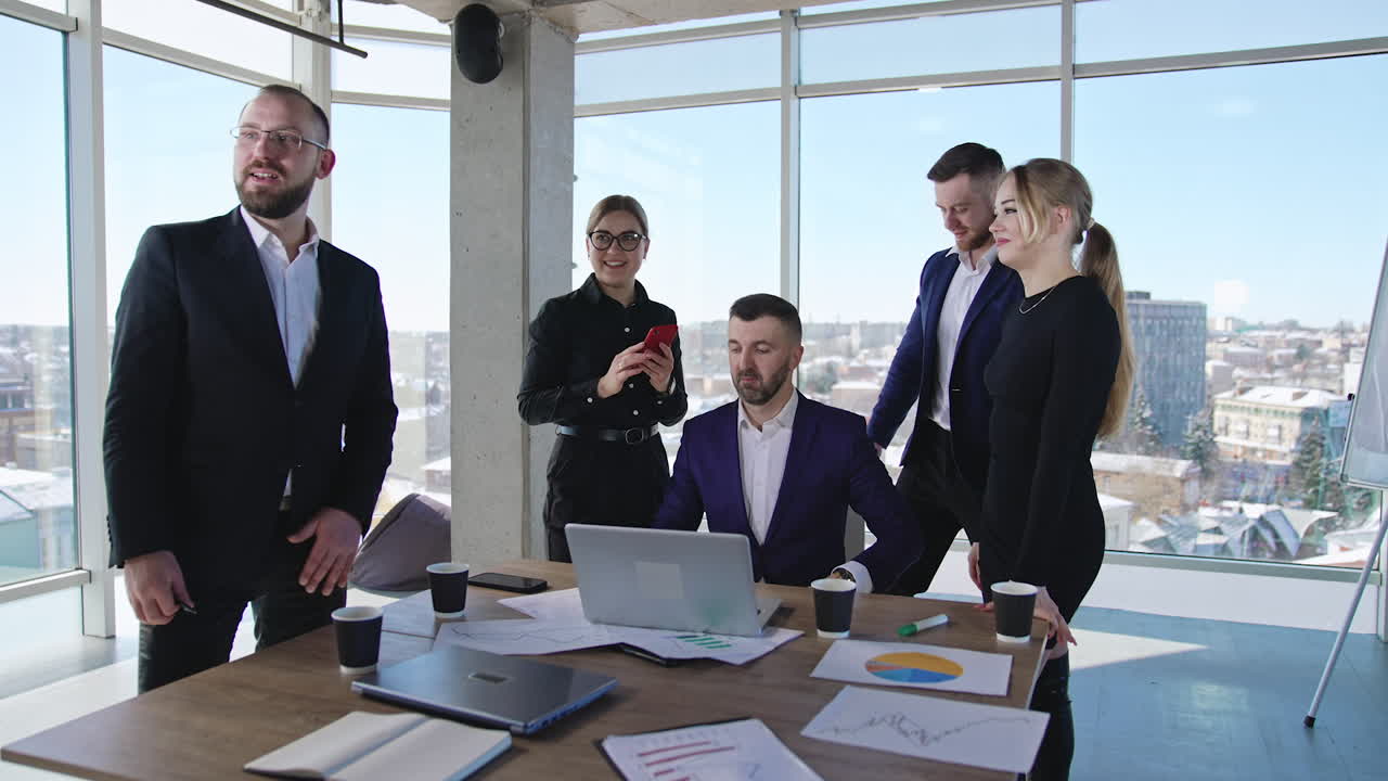 Business chief sits at the desk in front of laptop. Office team gathers around their leader. Panoramic windows with cityscape at backdrop.