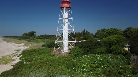 Beautiful-aerial-revealing-view-of-white-painted-steel-lighthouse-with-red-top-located-in-Pape,-Latvia-at-Baltic-sea-coastline-in-sunny-summer-day,-wide-angle-fast-ascending-drone-shot-moving-forward