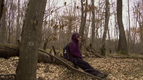 woman in purple jacket with backpack and water bottle sitting on a fallen tree in the forest