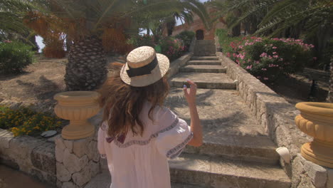 woman in a white dress and hat walking up stone steps in a garden with palm trees and pink flowers