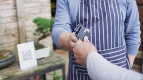 Caucasian-male-gardener-selling-plants,-using-contactless-payments-at-garden-center