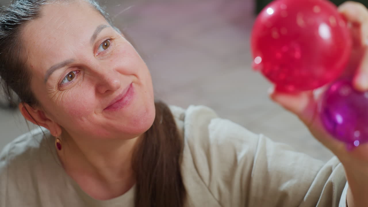 Aged woman smiling warmly while carefully examining colorful red and purple ornaments held in hand, surrounded by festive indoor setting filled with holiday spirit and Christmas decoration elements