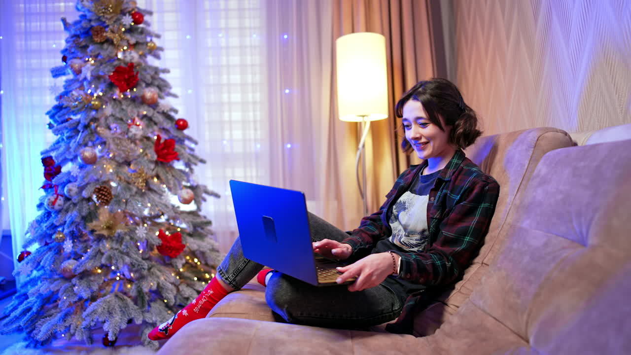 Happy brunette in checkered shirt and jeans sits on the sofa using her laptop. Girl doing online shopping for Christmas season.