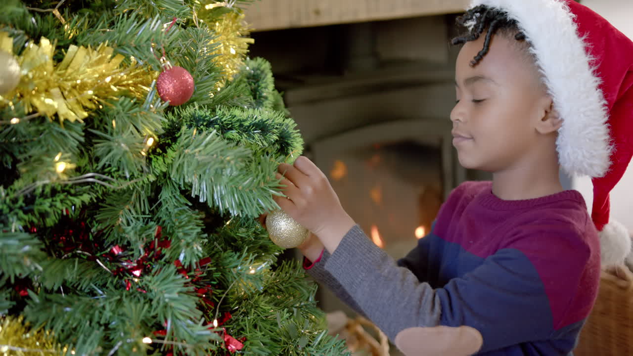 Happy african american boy in christmas hat decorating christmas tree, slow motion