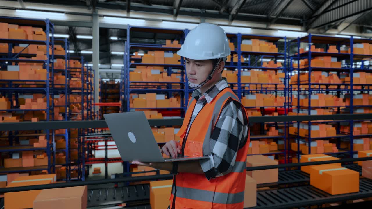 vista lateral de un ingeniero masculino asiático con casco de seguridad trabajando en una computadora portátil mientras está de pie en el almacén con estantes llenos de productos de entrega