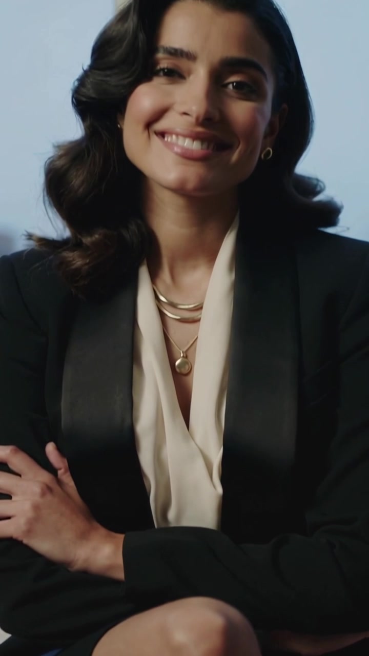 Video verticale: Zoom indietro che mostra una donna che si sposta, sorridendo alla finestra, in blazer e con collane d'oro.