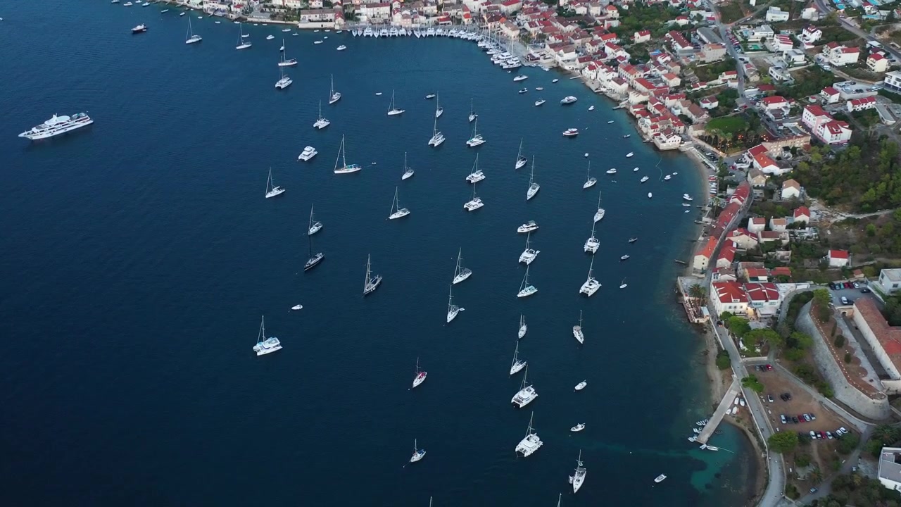 Yacht a vela e catamarani ancorati nella baia del porto di Vis, in Croazia, con banchine in pietra e case dai tetti rossi che si estendono lungo la costa al tramonto. Ripresa aerea grandangolare da drone