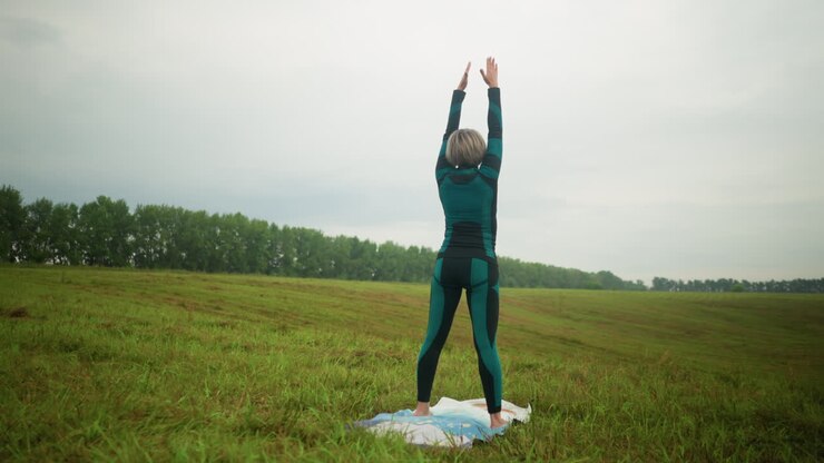 Back view of lady with hands raised up on yoga mat, slowly lowering them down, bending close to her knees, folding her arms, and twisting left and right, in a vast grassy field under a cloudy sky
