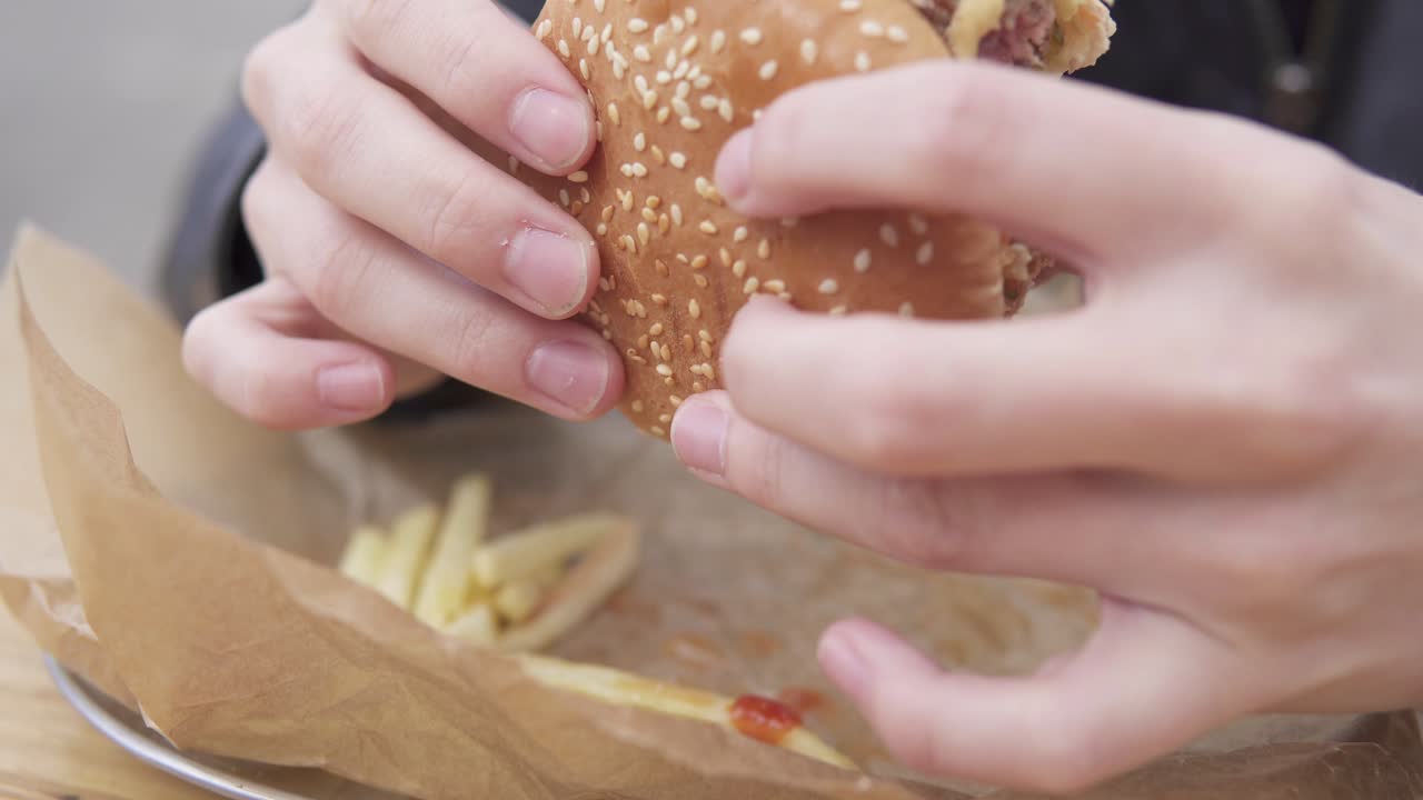 Closeup view of man's hands holding big tasty burger. Man with beard in the street cafe biting tasty big burger with cheese. French fries on the plate. Shot in 4k