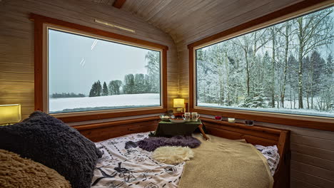 inside view of bed in corner of rustic wood cabin with large windows with view of snow winter landscape forest outside