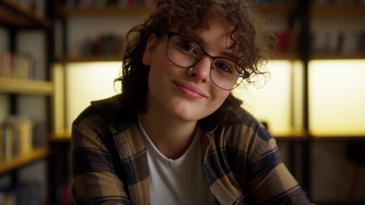 Contemplation of a happy girl student with curly hair and piercing who sits at a table in the library among books