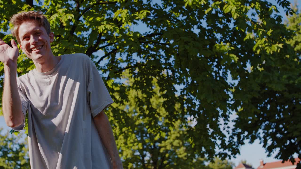 A cheerful man waves at the camera, surrounded by lush green trees