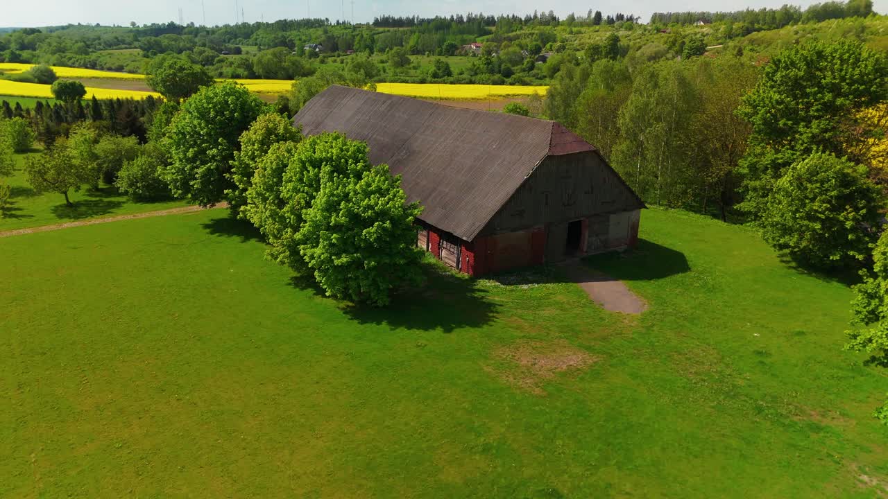 Luftfoto af en rustik lade omgivet af frodige grønne marker, træer og fjerne gule afgrødemarker.