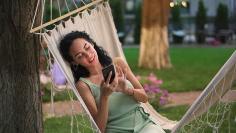 Portrait-Of-a-woman-relaxing-in-a-hammock-outdoors,-surfing-internet-in-her-smartphone,-clicks-on-the-screen-and-smiling