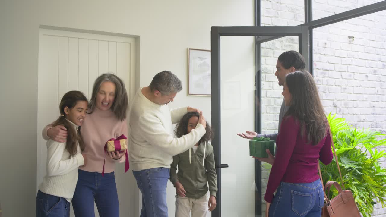 Senior man opening door at porch welcoming kids running hugging grandparents as adults handing gift