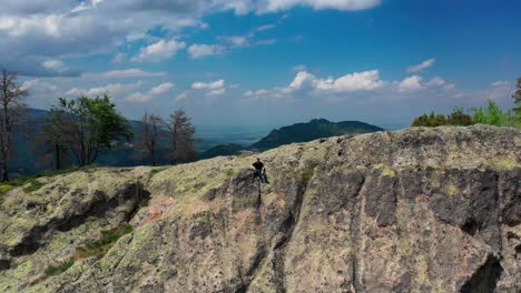 pull back shot of a man sitting on the side of a cliff revealing mountains, trees and cloudy blue sky