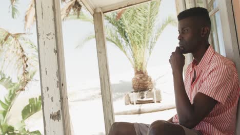 hombre afroamericano pensativo sentado en el porche de una casa de playa de madera mirando hacia otro lado, en cámara lenta