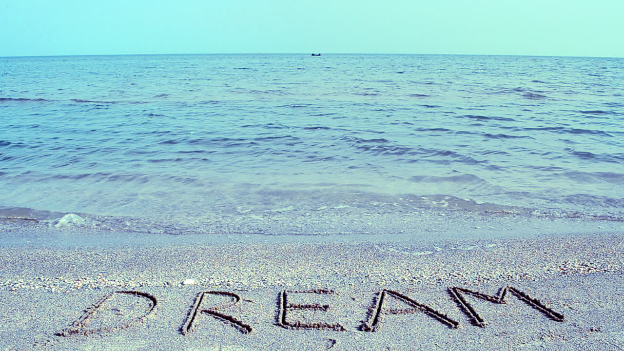 Scritta 'Dream' sulla sabbia della spiaggia, con l'onda. Riprese sulla spiaggia.