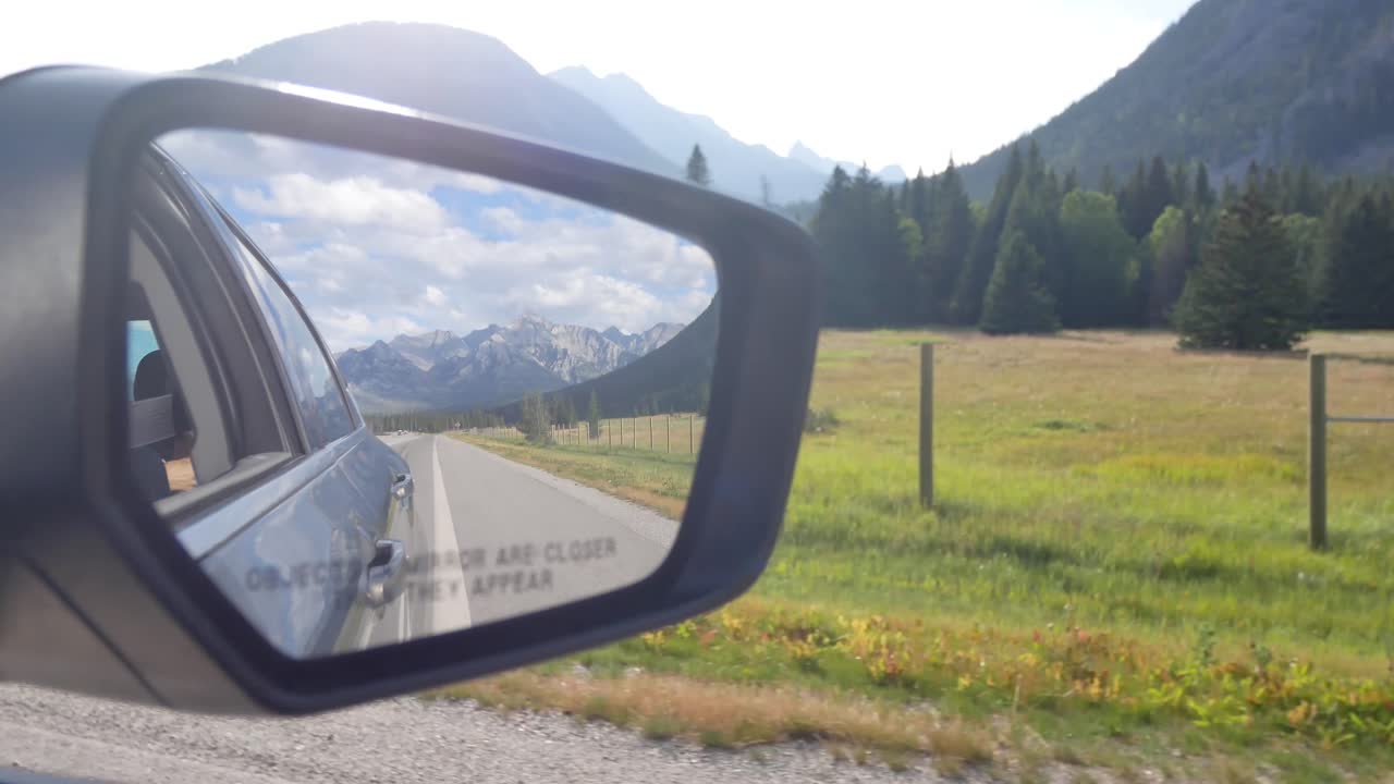 Landscape view of the side mirror`s car while moving with reflection view of the highway and surrounding with green pine tree forest and rockies mountain in Alberta,Canada