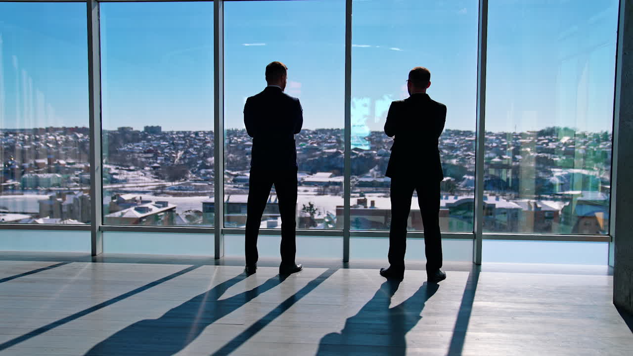 Office workers wearing suits talking indoors. Two businessmen standing in office and looking out of the windows. Rear view.