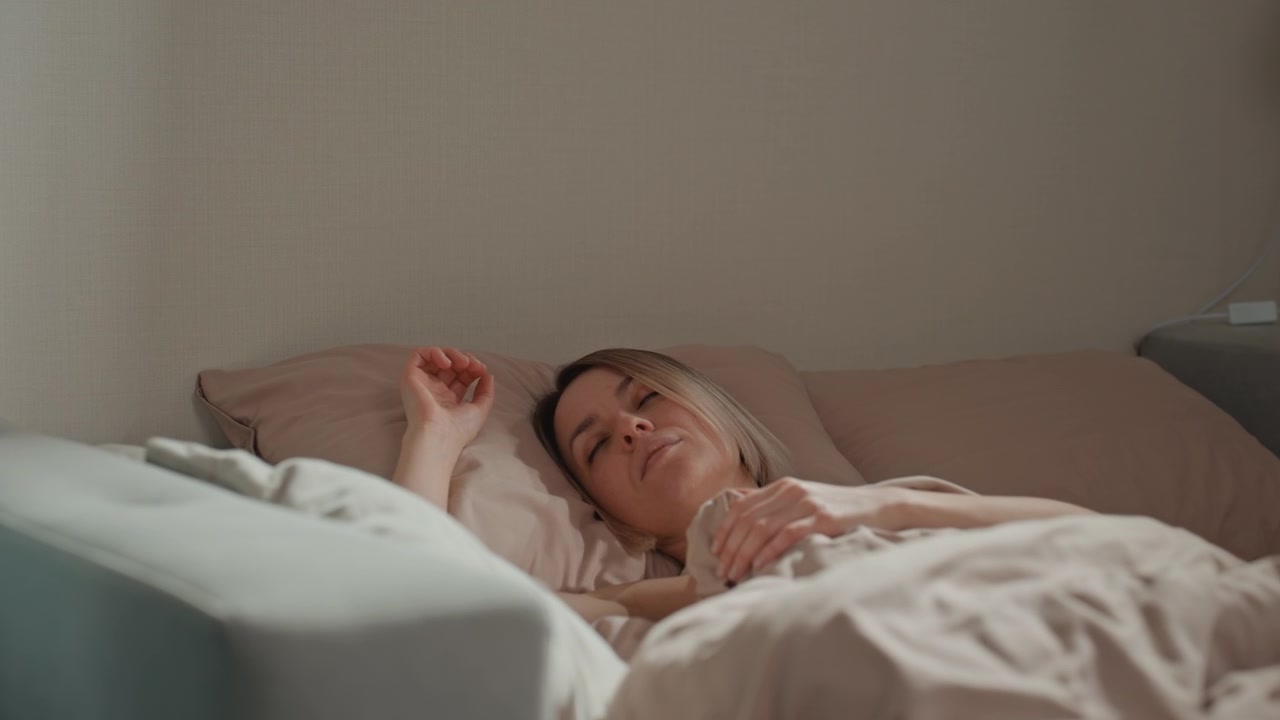 Kvinde hviler i seng, blødt morgenlys og neutralt sengetøj, nærbillede viser langsom vejrtrækning, hånd over pude, subtilt udtryk af eftertænksomhed og træthed, minimalistisk soveværelse med dæmpede farver.