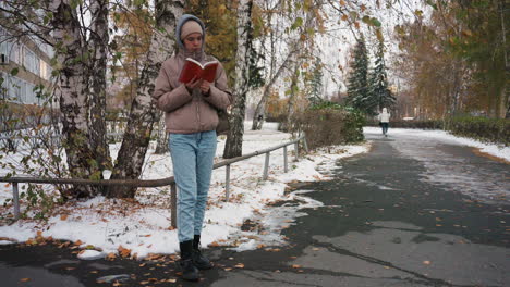 light skin woman dressed in winter clothing stands on snow-lined park walkway quietly reading book, surrounded by autumn trees, serene atmosphere with distant pedestrian in background