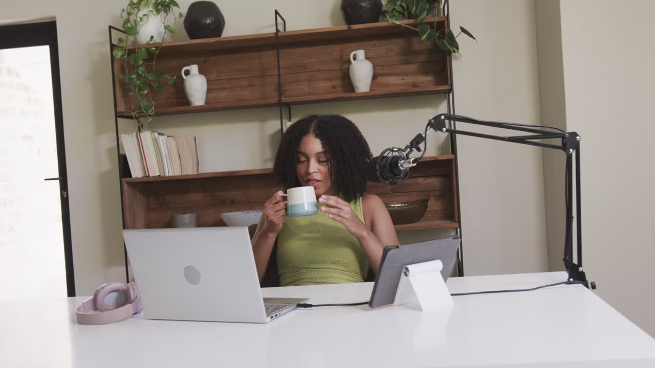 African American woman pausing typing and sipping mug, glancing at laptop, tablet at home office
