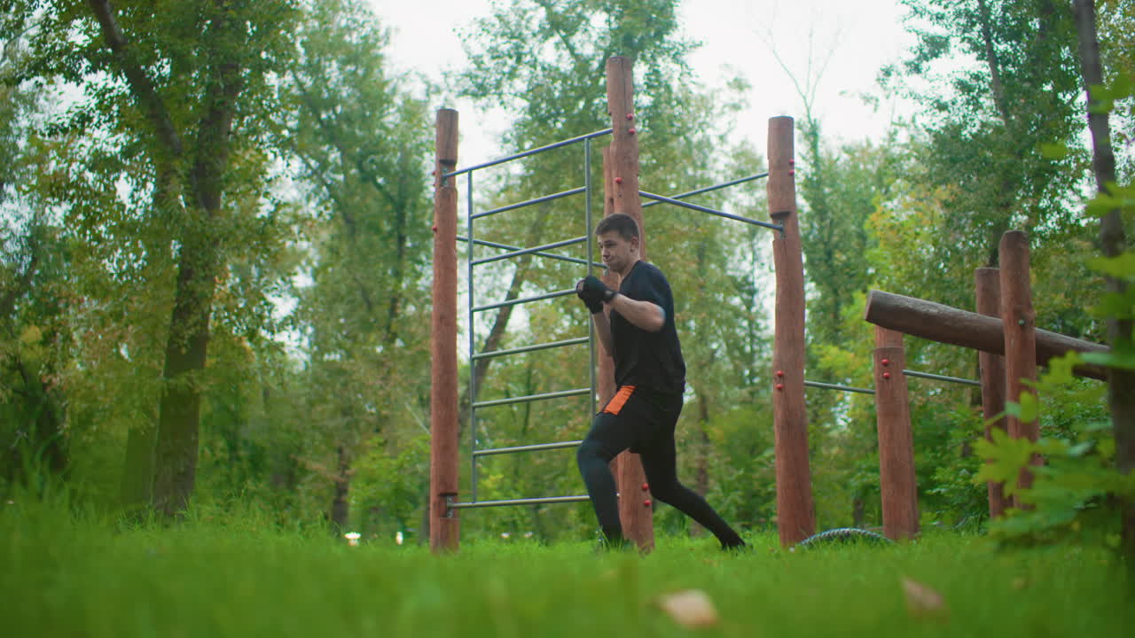 outdoor forest boxing training session with man shadowboxing near wooden obstacle course, focused footwork and controlled punches, green grass and tall trees, calm morning atmosphere, trainer role