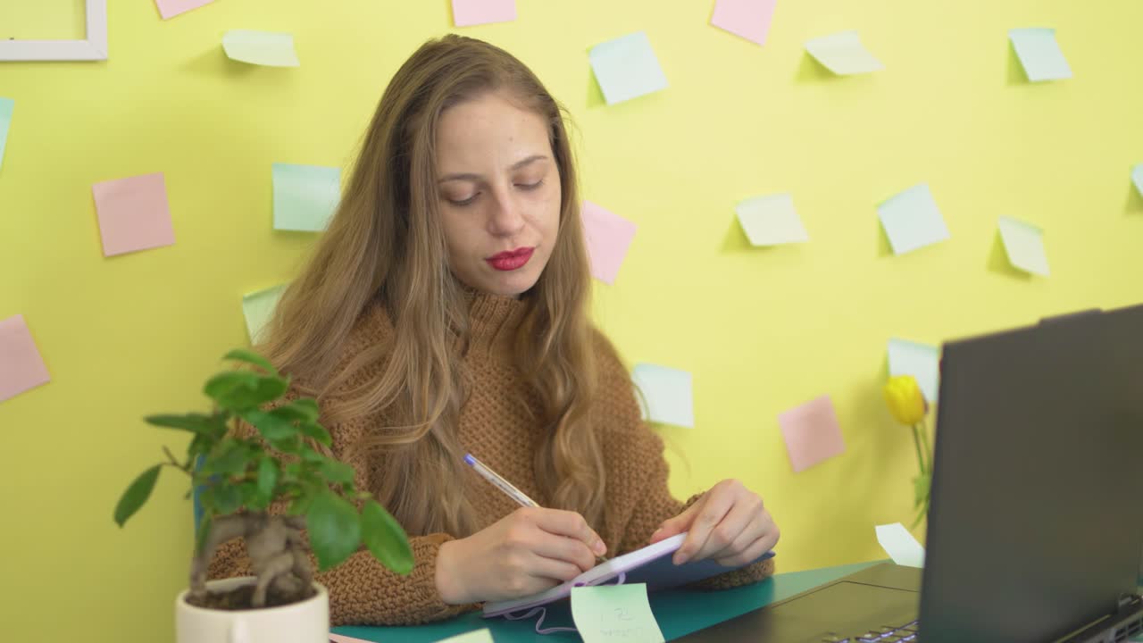 A young blonde Bulgarian woman writing an important note on a sticky note and sticking it to her laptop, set against a vibrant yellow backgroun